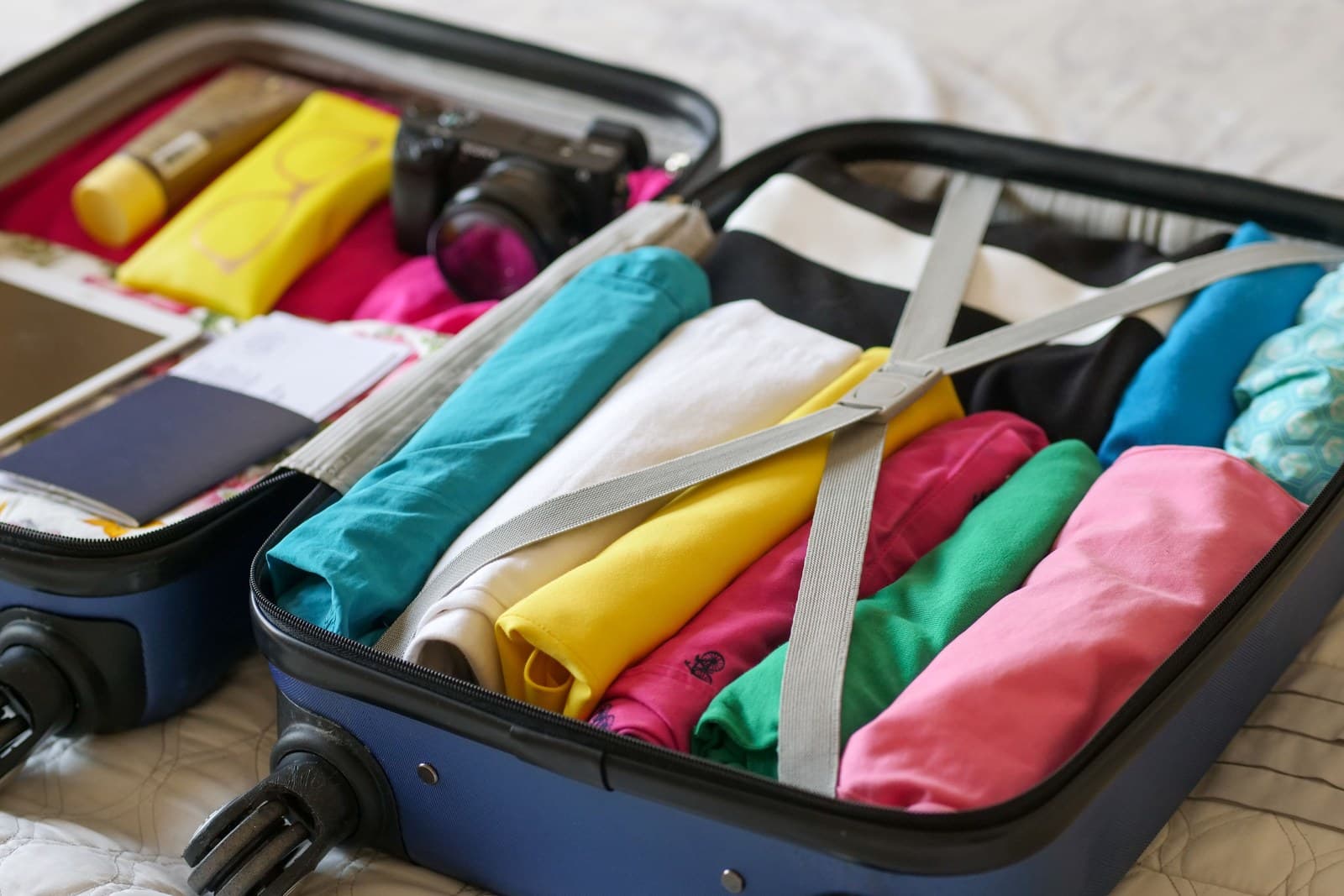 An open blue suitcase packed with rolled clothes, a camera and papers